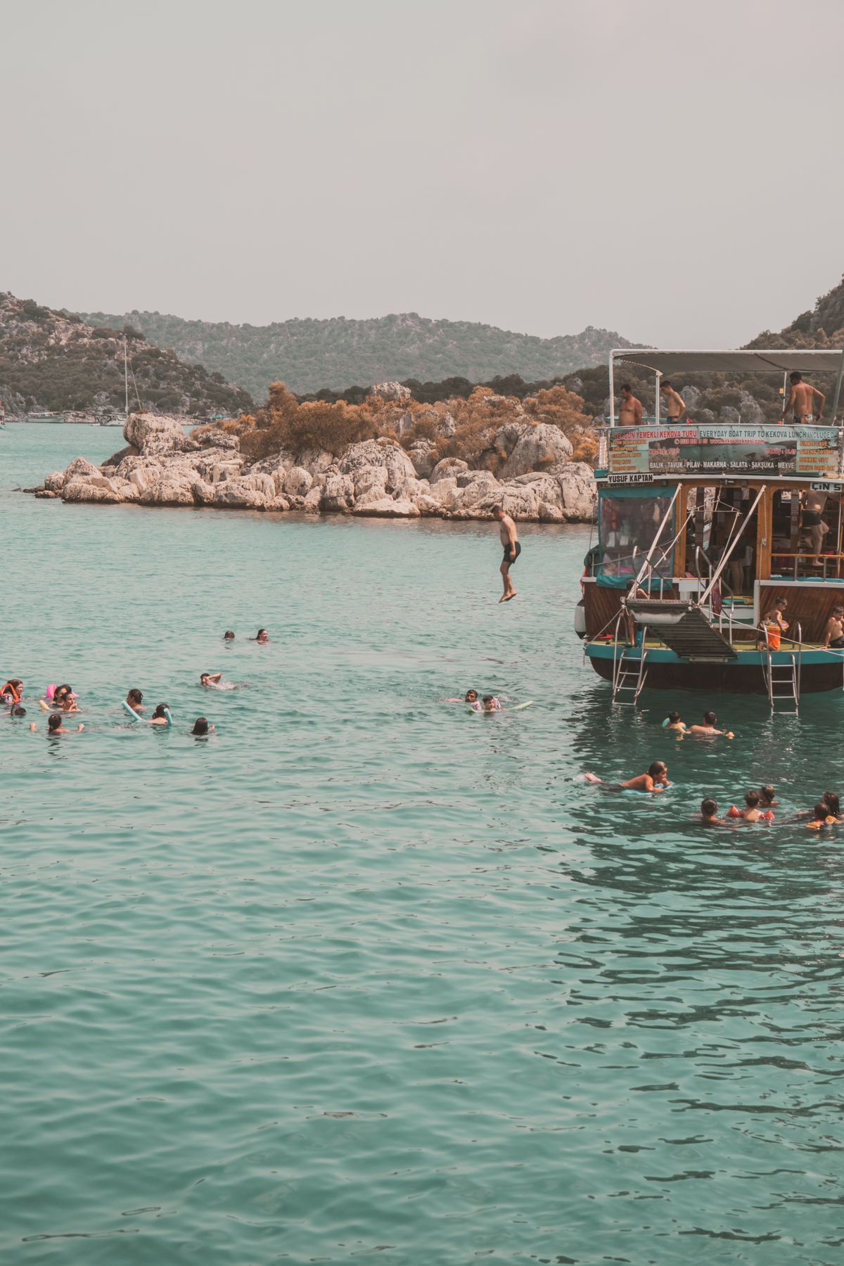 Guests swimming and diving from a boat near a rocky coastline on a sunny day